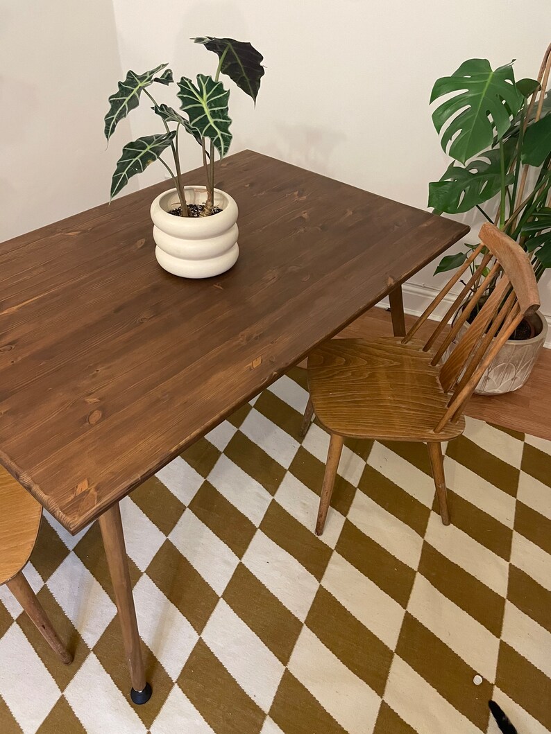 May include: A dark brown wooden dining table with a white ceramic planter and a green plant on top. The table has four legs and is sitting on a brown and white checkered rug. A wooden chair is pulled up to the table.