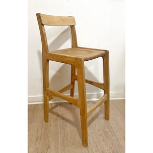 May include: A wooden bar stool with a simple, modern design. The stool is made of natural wood and has a light brown finish. It features a rectangular seat and a slightly curved backrest.