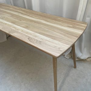 May include: A light-coloured wooden bench with a rectangular top and four angled legs. The bench has a natural wood grain pattern and a smooth, polished finish. The legs are slender and tapered, providing a stable base. The bench is approximately 152 cm long.