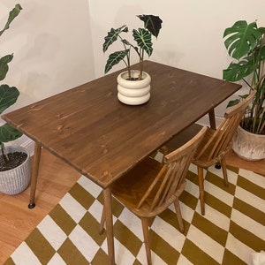 May include: A dark brown wooden dining table with a white ceramic planter and a green plant on top. The table has four legs and is set on a black and white chequered rug. Two wooden chairs are placed at the table.