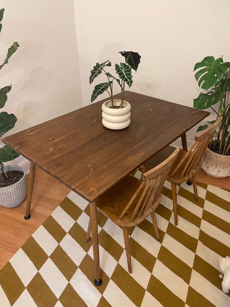 May include: A dark brown wooden dining table with a white ceramic planter and a green plant on top. The table has four legs and is set on a black and white chequered rug. Two wooden chairs are placed at the table.