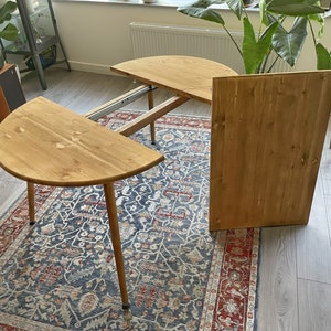 May include: A wooden table with a round, extendable top. The table is made of light brown wood and has a natural finish. The table is sitting on a rug with a blue and red pattern.