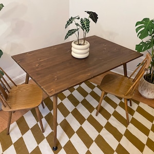 May include: A dark brown wooden dining table with a white and brown chequered rug underneath. Two wooden chairs are placed on either side of the table. A potted plant with green leaves sits on the table.