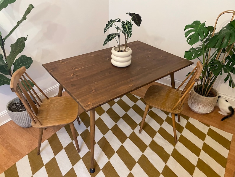 May include: A dark brown wooden dining table with a white and brown chequered rug underneath. Two wooden chairs are placed on either side of the table. A potted plant with green leaves sits on the table.