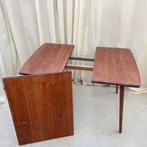 May include: A wooden dining table with a warm, reddish-brown finish. The table has a unique design with a leaf extension, allowing it to expand. The table is supported by slender, tapered legs. The table is approximately 152 cm long.