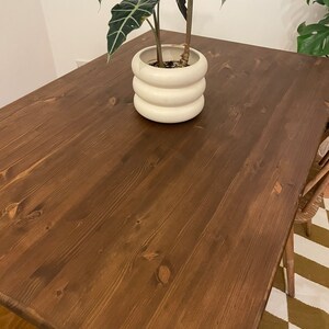 May include: A dark brown wooden table with a natural wood grain finish. A white ceramic planter with a plant is on the table.