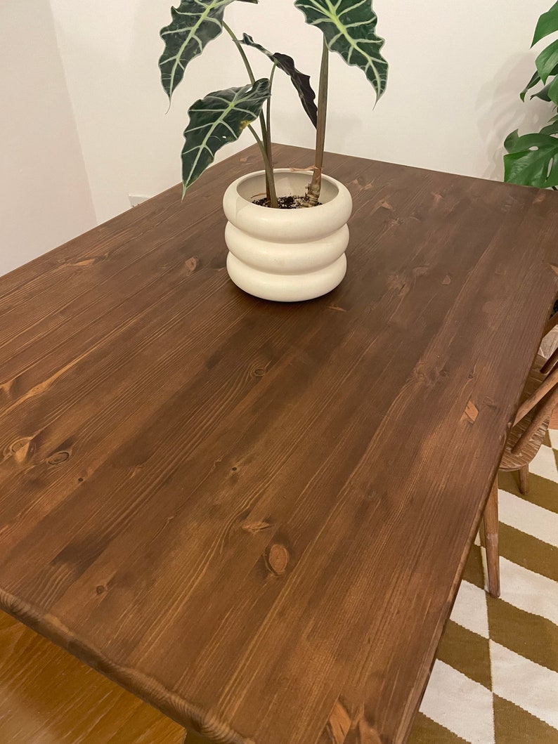 May include: A dark brown wooden table with a natural wood grain finish. A white ceramic planter with a plant is on the table.