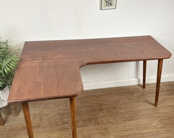 May include: An L-shaped wooden desk with a dark brown stained finish and a smooth surface. The desk features a curved corner design and is supported by four cylindrical wooden legs that extend straight down to the floor from the tabletop corners.