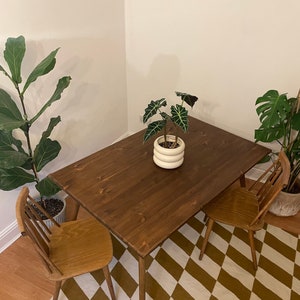 May include: A dark brown wooden dining table with two chairs. The table has a simple design with four legs and a rectangular top. The chairs are made of light brown wood and have a slatted back and seat. The table and chairs are set on a white and brown diamond patterned rug.