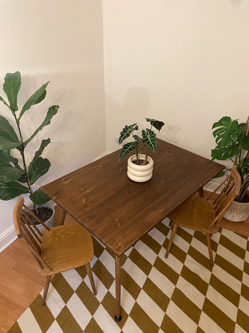 May include: A dark brown wooden dining table with two chairs. The table has a simple design with four legs and a rectangular top. The chairs are made of light brown wood and have a slatted back and seat. The table and chairs are set on a white and brown diamond patterned rug.