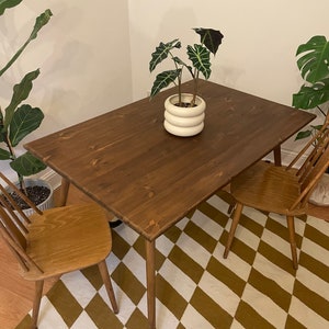 May include: A dark brown wooden table with a white ceramic planter and a green plant on top. The table has four legs and is sitting on a brown and white chequered rug. Two wooden chairs are placed on either side of the table.