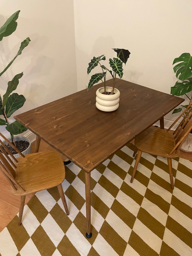 May include: A dark brown wooden table with a white ceramic planter and a green plant on top. The table has four legs and is sitting on a brown and white chequered rug. Two wooden chairs are placed on either side of the table.