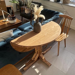 May include: A light brown wooden semi-circular drop-leaf table with a central pedestal base, accompanied by two matching wooden spindle-back chairs. A dark grey ceramic vase containing fluffy white pampas grass sits on the tabletop.