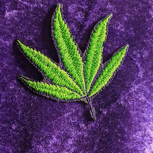 May include: A green embroidered marijuana leaf patch on a purple velvet background.