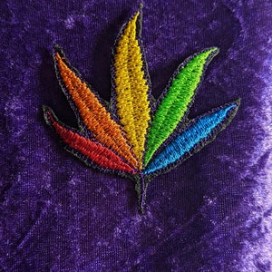 Puede incluir: Un parche bordado de hoja de marihuana de colores arcoíris. La hoja está bordeada de negro y tiene un tallo verde oscuro.