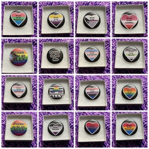 May include: A set of 16 button pins with various pride flags and messages. The pins are in white boxes with a purple background. The pins are all heart-shaped and feature different pride flags and messages such as "Proud to be an Ally", "Proud to be Me", "Proud to be Transgender", "Proud to be Intersexual", "Proud to be Straight Ally", "Proud to be Genderfluid", "Proud to be Asexual", "Proud to be Demisexual", "Proud to be Polysexual", "Proud to be Non-Binary", "Proud to be Lesbian", "Proud to be Agender", "Proud to be Gay", and "Proud to be Pansexual".