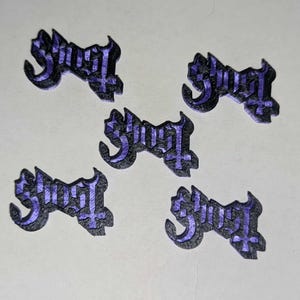 Op de afbeelding: Vijf zwarte en paarse Ghost logo patches. Het logo heeft gestileerde tekst met een kruissymbool. De patches zijn gerangschikt op een lichtgekleurd oppervlak.
