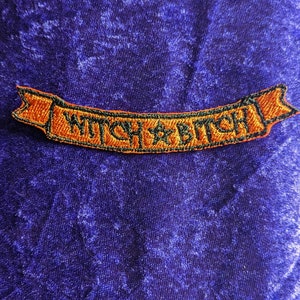 May include: Orange and black embroidered patch with the text "Witch Bitch" on a purple velvet background.