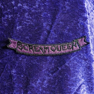 Peut inclure: Un écusson brodé violet avec le texte "SCREAM QUEEN" en lettres vertes. L'écusson est en forme de bannière avec des extrémités pointues et une bordure noire. Le fond est un tissu violet foncé texturé.