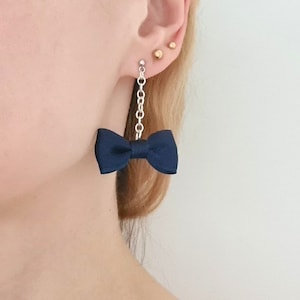 Puede incluir: Un par de pendientes colgantes con un lazo azul marino y una cadena plateada. Los pendientes son usados por una persona con cabello rubio.