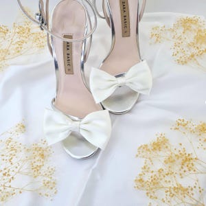 Puede incluir: Un par de sandalias plateadas de tacón alto con lazos de satén blanco. Las sandalias tienen tiras para el tobillo y la marca "ZARA BASIC" está impresa en las tiras.