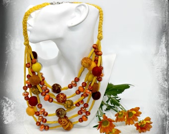 May include: A multi-strand necklace with felt balls in shades of orange, yellow, and brown. The necklace is made with a braided cord and is displayed on a white mannequin.