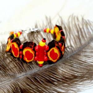 May include: A red, yellow, and black felt bracelet with a silver clasp. The bracelet is on a brown feather.