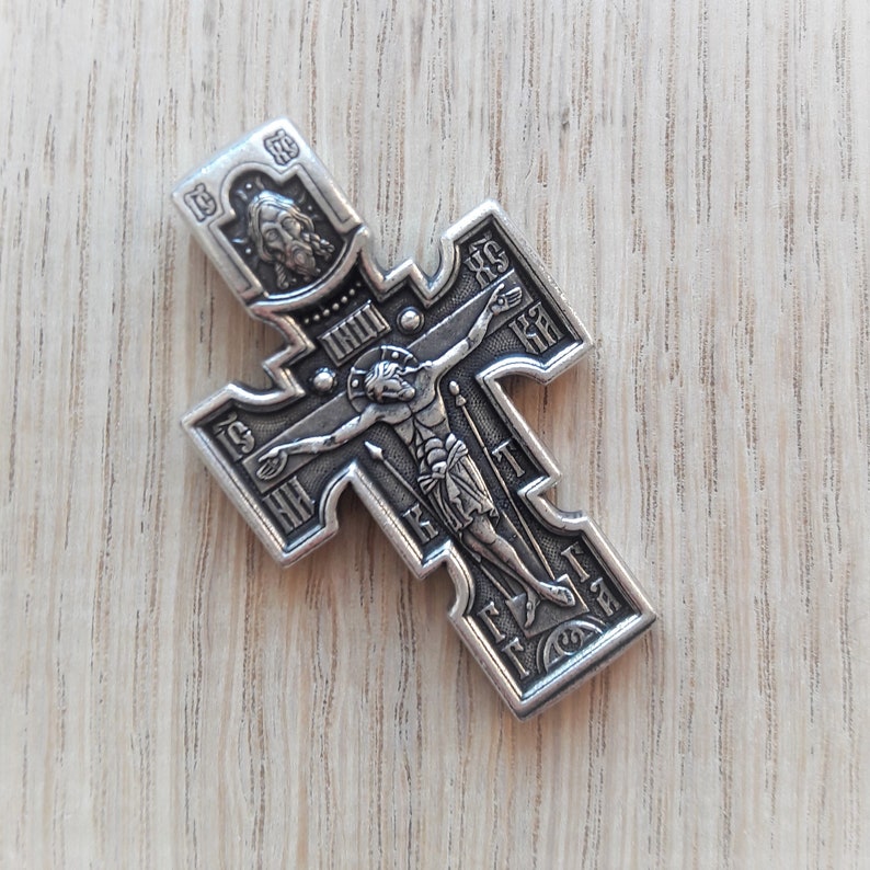 K&ouml;nnte beinhalten: Ein silberfarbener Kreuzanh&auml;nger mit einem komplizierten Design, das eine Figur von Jesus Christus am Kreuz zeigt. Das Kreuz hat kyrillische Schriftzeichen an den R&auml;ndern.