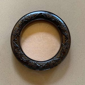 May include: A round, dark brown picture frame with a floral design. The frame has a circular opening and a light brown backing. The frame's border is adorned with detailed floral patterns, adding a decorative touch to the design.