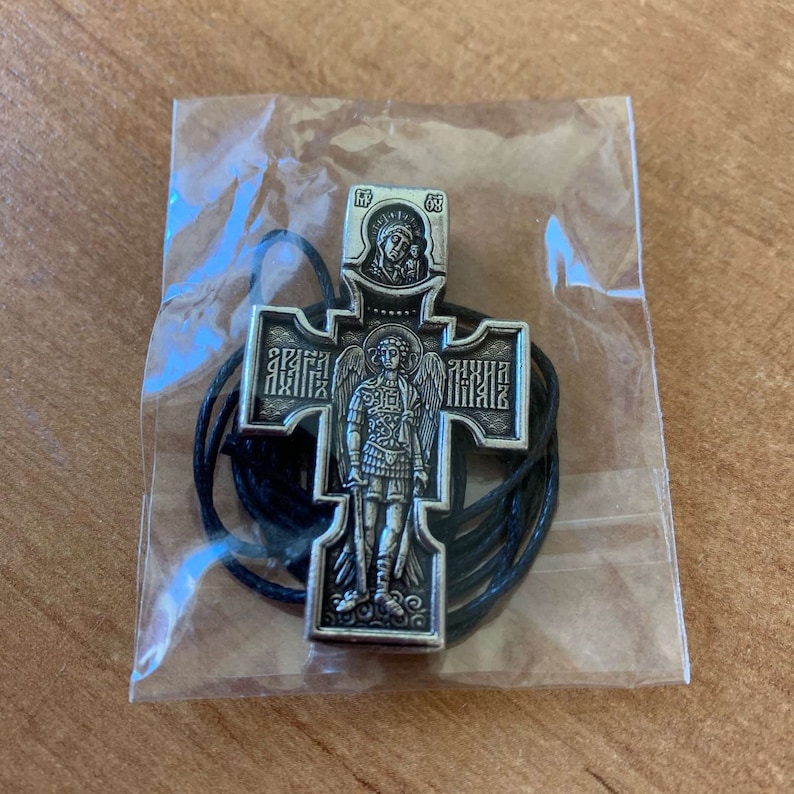 K&ouml;nnte beinhalten: Ein silberfarbener Kreuzanh&auml;nger mit einem schwarzen Band. Das Kreuz zeigt ein Bild eines Engels mit Fl&uuml;geln und einem Schwert sowie die Inschrift "СВЯТАЯ ТРОИЦА" in kyrillischer Schrift. Oben auf dem Kreuz befindet sich ein kleines Bild der Jungfrau Maria und des Kindes.