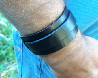Könnte beinhalten: Schwarzes Lederarmband. Das Armband besteht aus drei übereinanderliegenden Bändern aus schwarzem Leder. Das Armband wird an einem Handgelenk getragen, mit Jeans und einem blauen Hemd im Hintergrund.