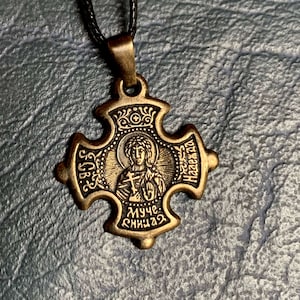 Może przedstawiać: Złoty metalowy wisiorek w kształcie krzyża z przedstawieniem kobiety w środku. Wisiorek ma cyrylicę wokół krawędzi, w tym "Муче" i "Назарека".