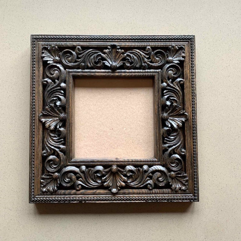 May include: A dark brown wooden picture frame with an ornate, carved design. The frame is square and has a plain interior.