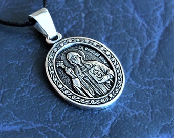 May include: Silver pendant necklace with a religious icon of Saint Barbara. The icon is oval shaped and features a detailed image of Saint Barbara holding a book and a cross. The pendant is on a black cord.