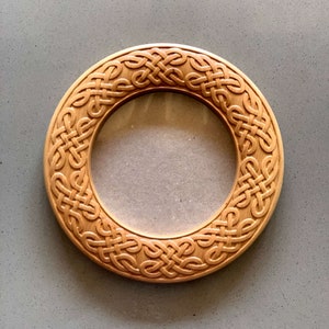 May include: A round wooden frame with a Celtic knot design carved into the surface. The frame is made of light brown wood and has a smooth finish.