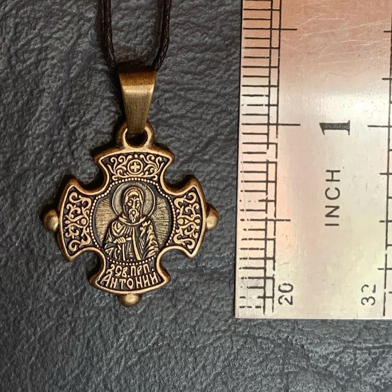 May include: A gold-toned metal pendant in the shape of a cross with an image of a saint inside a circle. The pendant is about 1 inch in diameter and has a black cord attached to it. The pendant has the text "298. прп. АНТОНИН" on the bottom.