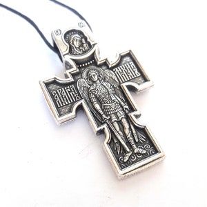 K&ouml;nnte beinhalten: Silberner Anh&auml;nger in Form eines Kreuzes mit einem Engel und einer religi&ouml;sen Inschrift. Das Kreuz befindet sich an einer schwarzen Schnur.