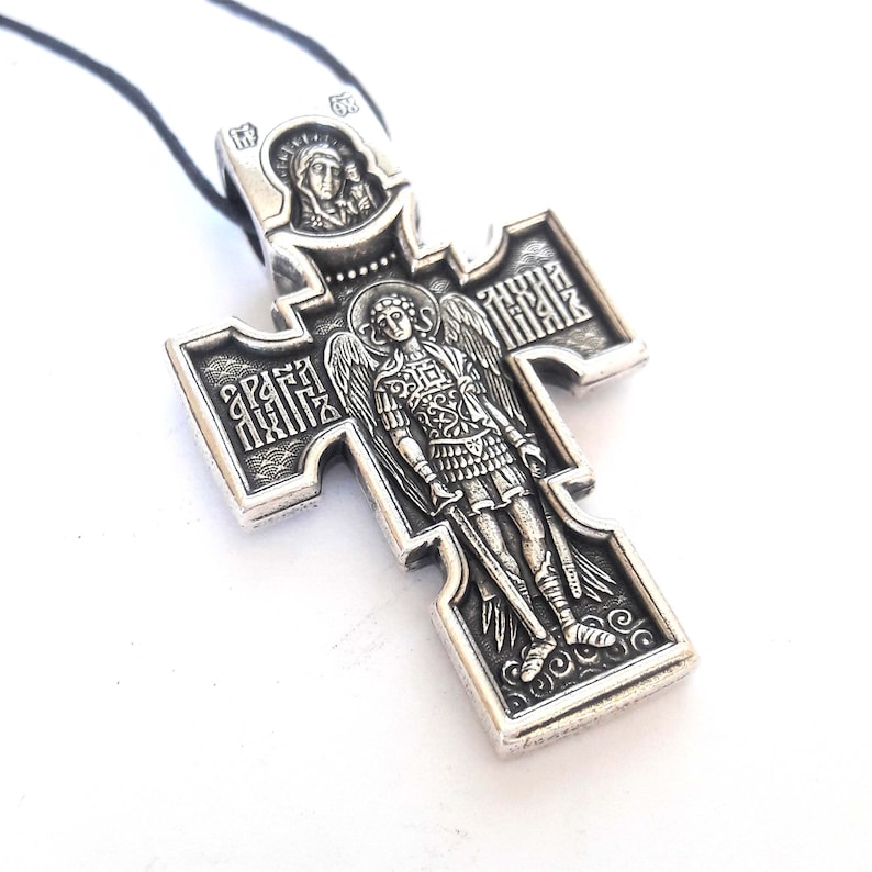 K&ouml;nnte beinhalten: Silberner Anh&auml;nger in Form eines Kreuzes mit einem Engel und einer religi&ouml;sen Inschrift. Das Kreuz befindet sich an einer schwarzen Schnur.