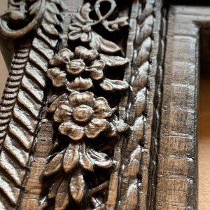 May include: Close-up of a dark brown wooden picture frame with intricate carvings of flowers and leaves. The frame has a distressed finish and a rope-like design on the right side.