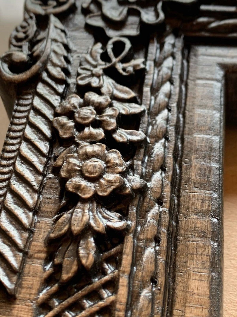 May include: Close-up of a dark brown wooden picture frame with intricate carvings of flowers and leaves. The frame has a distressed finish and a rope-like design on the right side.