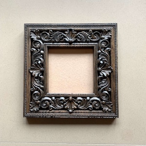 May include: A dark brown wooden picture frame with an ornate, carved design. The frame is square shaped and has a raised edge.