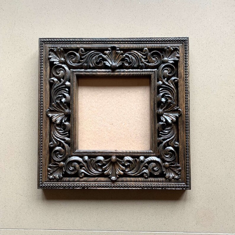 May include: A dark brown wooden picture frame with an ornate, carved design. The frame is square shaped and has a raised edge.