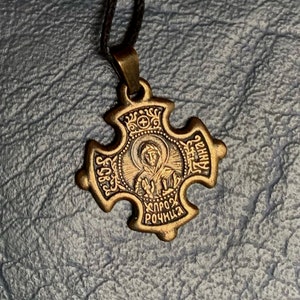 May include: A gold-toned metal pendant in the shape of a cross with a depiction of a religious icon in the center. The icon is surrounded by a halo and the words "Спроз Рочница" are inscribed below it. The cross has four arms, each with a different inscription: "Св", "Ангел", "Св", and "Ангел".