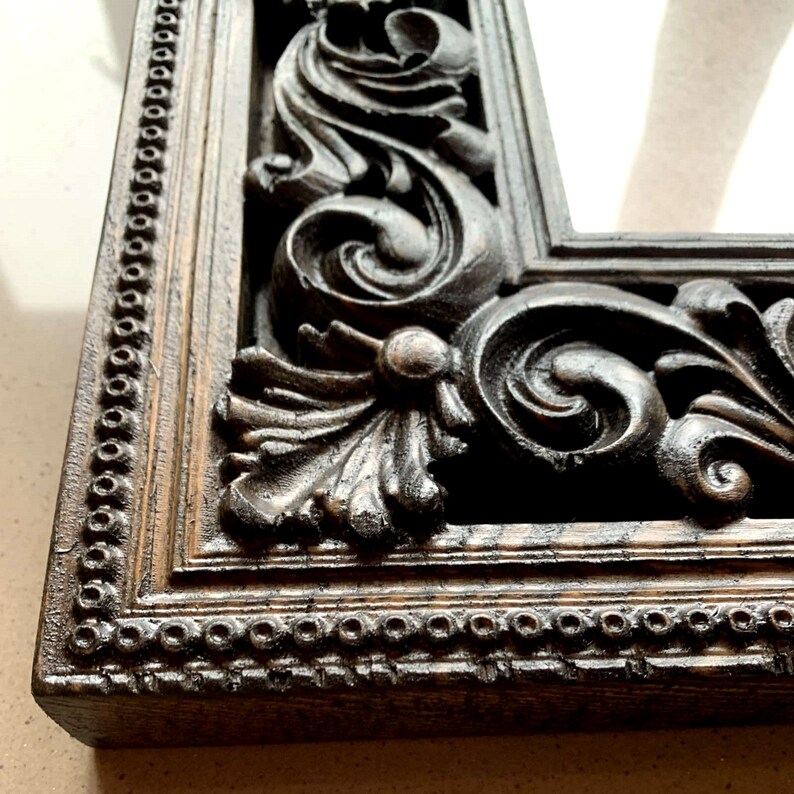 May include: A close-up of a dark brown ornate mirror frame with intricate carvings. The frame has a beaded edge and a distressed finish.