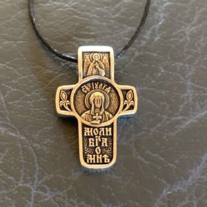 Puede incluir: Un colgante de cruz de plata con un cordón negro. La cruz presenta una imagen de una mujer con un halo y el texto "СВ. ИОЛИЯ" encima de ella. El texto "ЯОЛН БГА О ЖНГ" está inscrito debajo de la imagen.