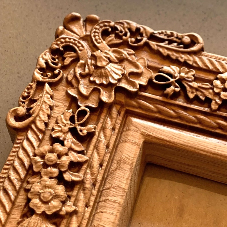 May include: A close-up of a wooden picture frame with intricate carvings of flowers and leaves. The frame is a light brown color and has a smooth, polished finish.