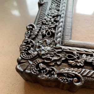 May include: Close-up of a dark gray ornate picture frame with intricate floral carvings. The frame is made of wood and has a distressed finish.
