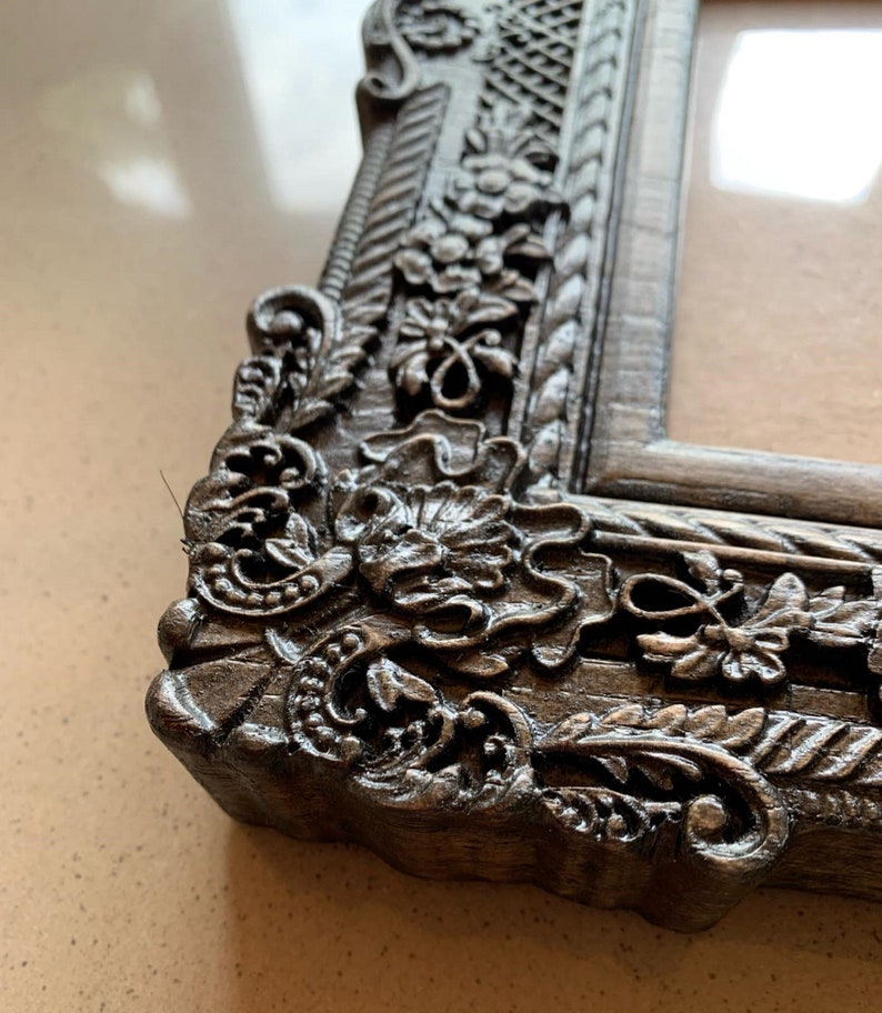 May include: Close-up of a dark gray ornate picture frame with intricate floral carvings. The frame is made of wood and has a distressed finish.