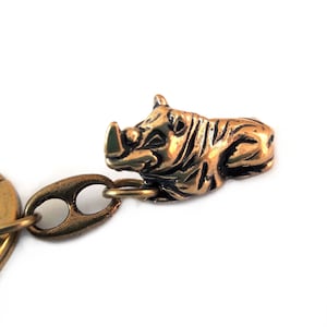 May include: A gold-toned keychain featuring a detailed rhinoceros figurine. The rhino is lying down with its head turned to the side. The keychain is made of metal and has a ring for attaching keys.
