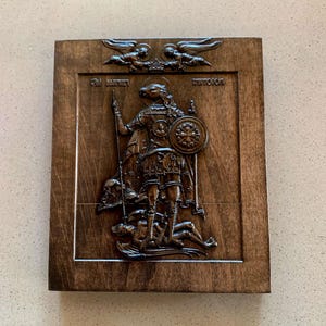 May include: A dark brown wooden plaque with a carved relief depicting a figure in armor holding a spear and shield, standing over a defeated figure. Two angels are carved at the top. The plaque has a framed border.